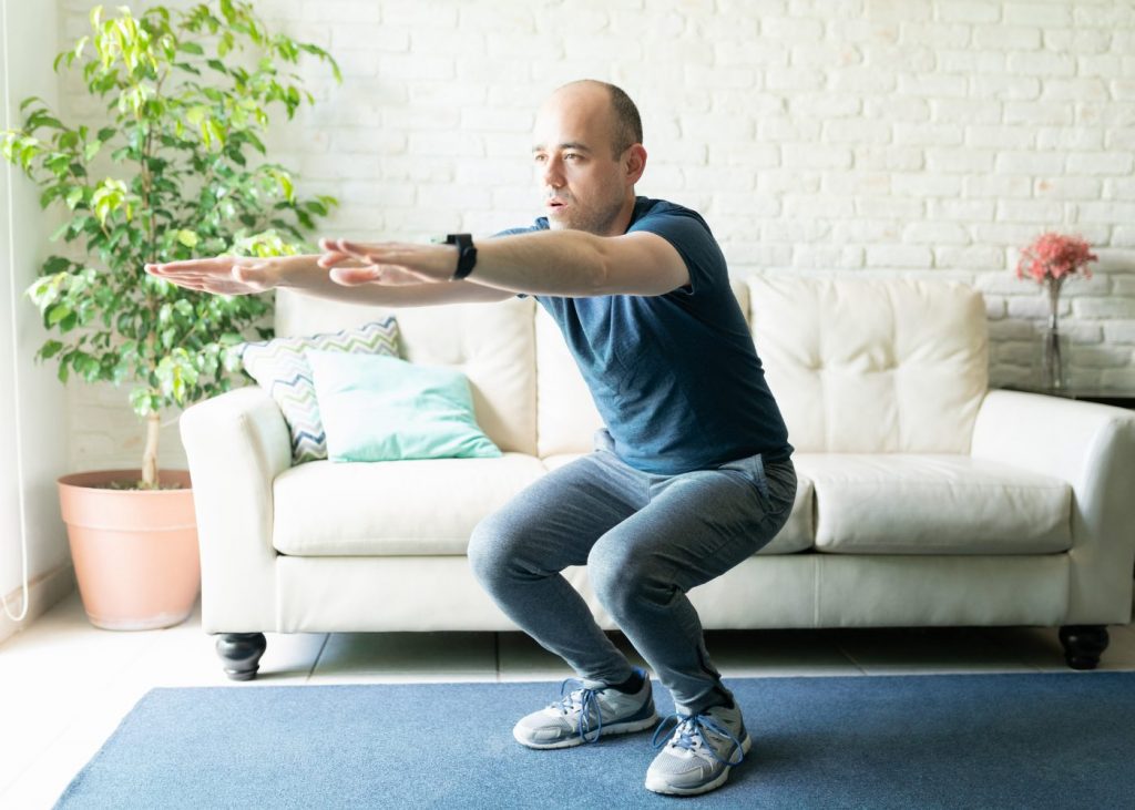 Mann führt Kniebeuge im Wohnzimmer aus – Übung zur Kräftigung von Knie, Hüfte und Beinmuskulatur bei physiotherapeutischer Behandlung